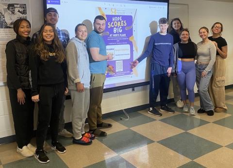 Westfield State University class of Advanced Public Relations pose in front of a poster design for “Stick Together with Rick’s Place Where Hope Scores Big,” campaign.