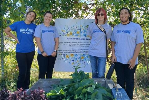 Four students stand in front of garden plants and a sign for Grandmothers' Garden