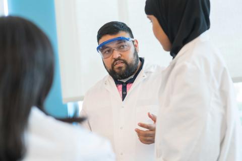 Roderico Acevedo teaches in a lab coat and goggles while two students also in lab coats look on