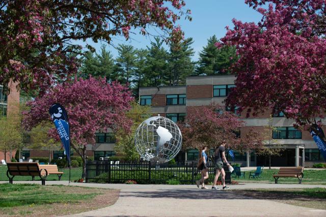 Two students walk in front of the campus globe surrounded by pink flowering trees on a bright spring day.